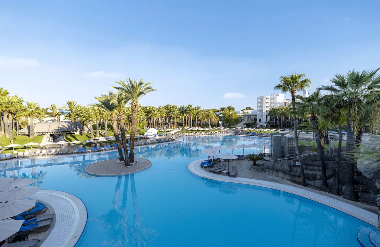 The main pool at Monachus Family Resort Sorgun, a large freeform pool with a palm island at its centre, ringed by sun loungers, parasols and palms.