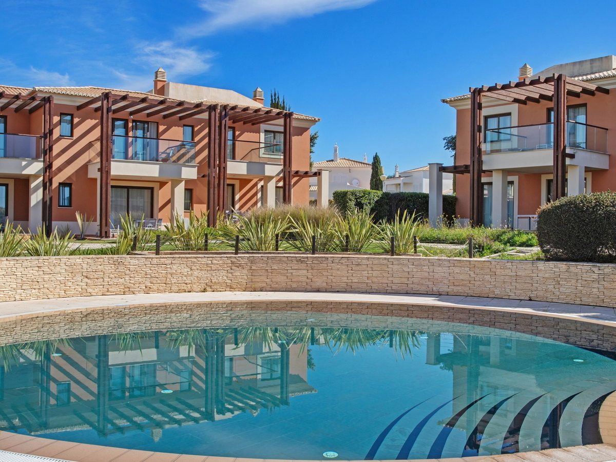 A small round pool with wide steps, set below a stone retaining wall with two-storey terracotta villas and pergolas behind.