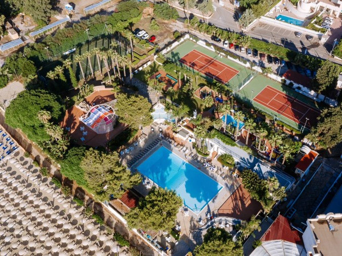 The pool area from the air, with a rectangular blue pool, a smaller pool, planted grounds, tennis courts and rows of beach parasols alongside.