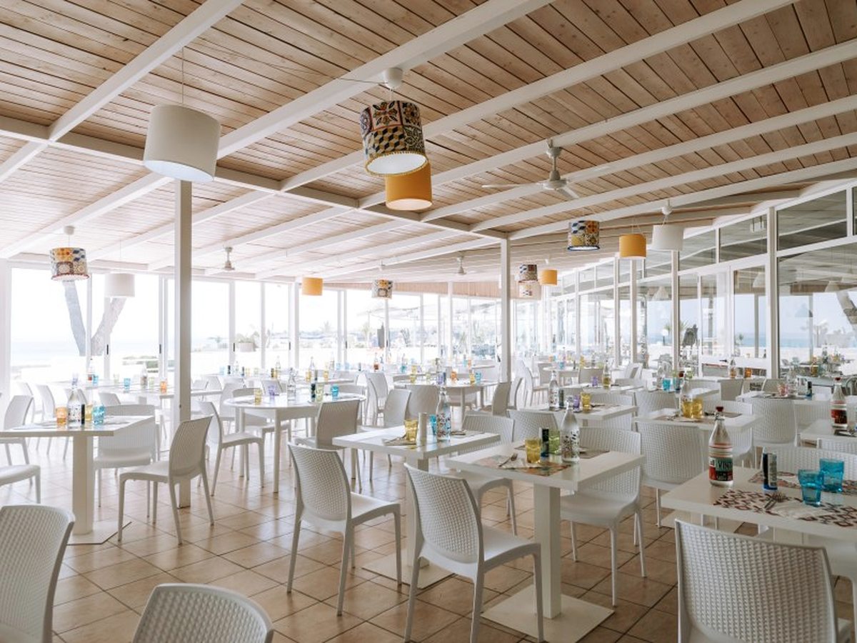 A large open-sided dining room under a pale timber ceiling with ceiling fans, filled with white tables and chairs, with the sea beyond.