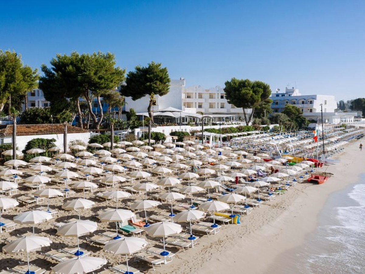 Rows of cream parasols and sun loungers on the sand in front of the resort's white buildings and pine trees.