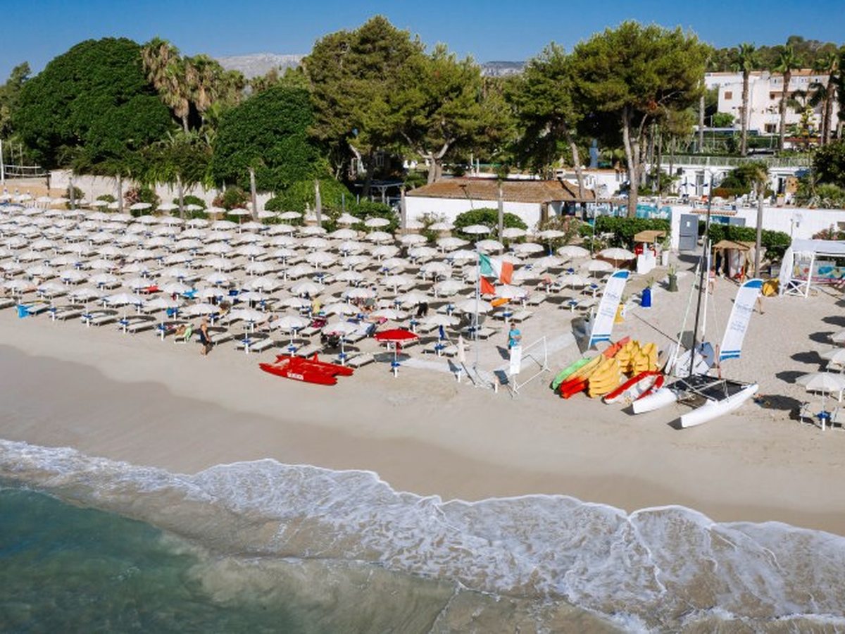 Kayaks, a catamaran and pedalos drawn up on the sand at the end of the rows of parasols, with the resort's trees and buildings behind.