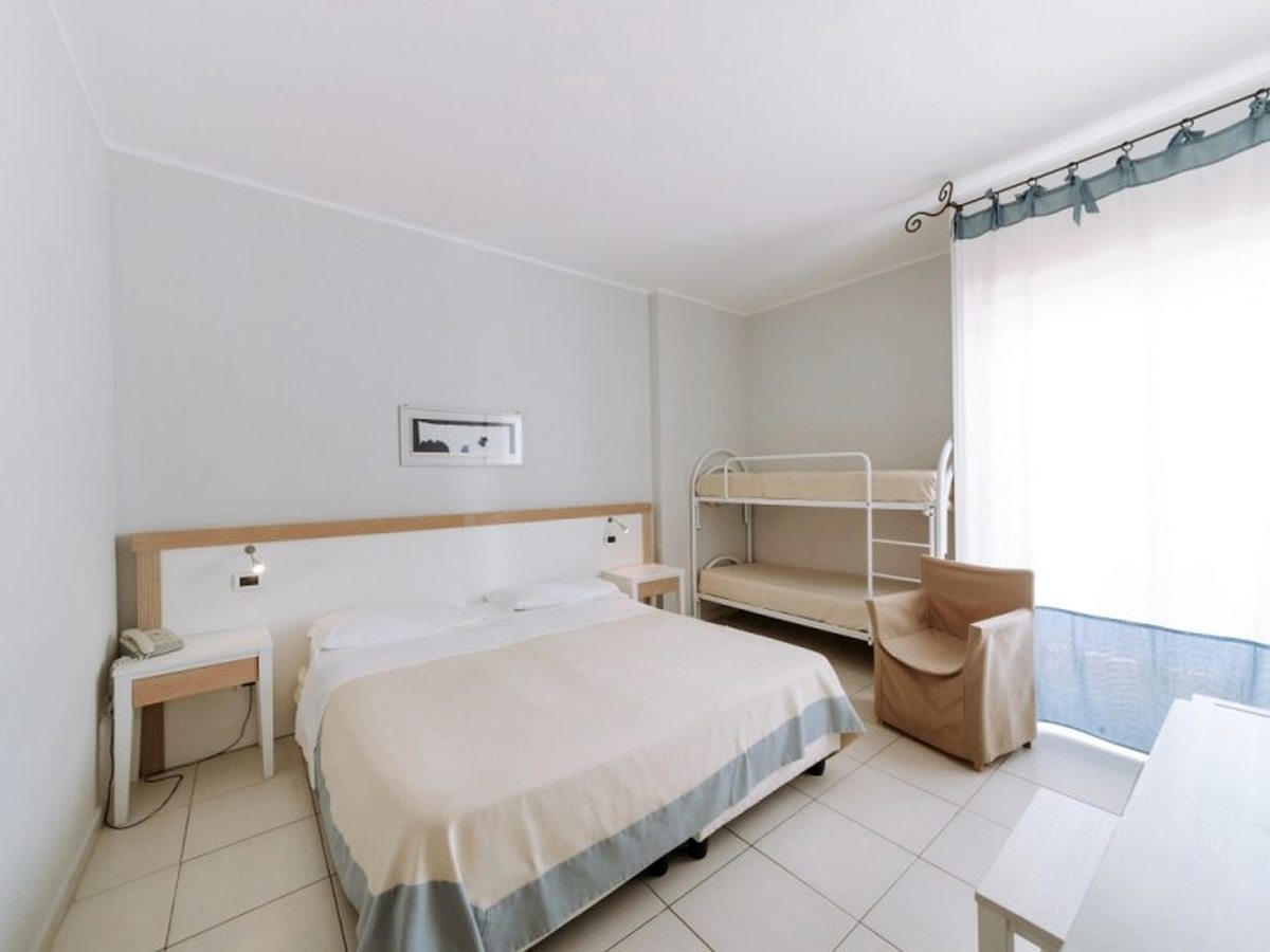 A room with a double bed and a white metal bunk bed beside a curtained window, with an armchair and a tiled floor.
