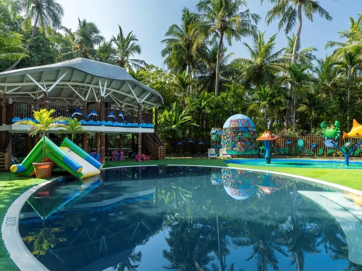 A shallow children's pool with a curved edge, small slides and water-spray play pieces, set on a lawn among palm trees.