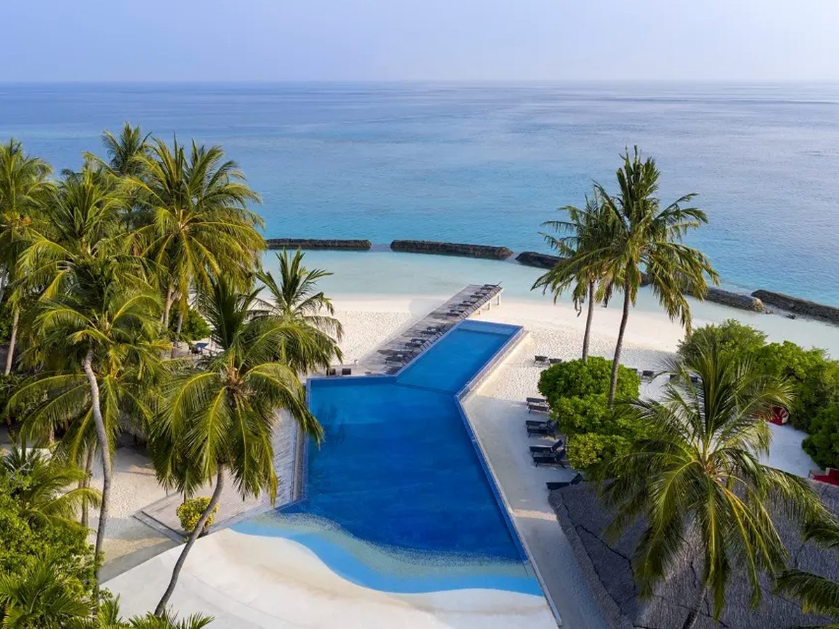 An L-shaped pool set into white sand behind a line of palms, with sun loungers along one side, a row of loungers on a jetty reaching to the beach and the pale sea beyond.