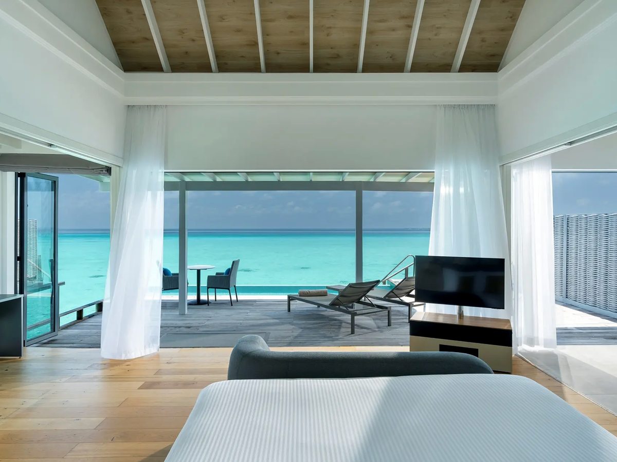 The inside of an overwater villa with a pale timber ceiling and white curtains, opening across a deck with sun loungers and a small infinity pool to the lagoon.