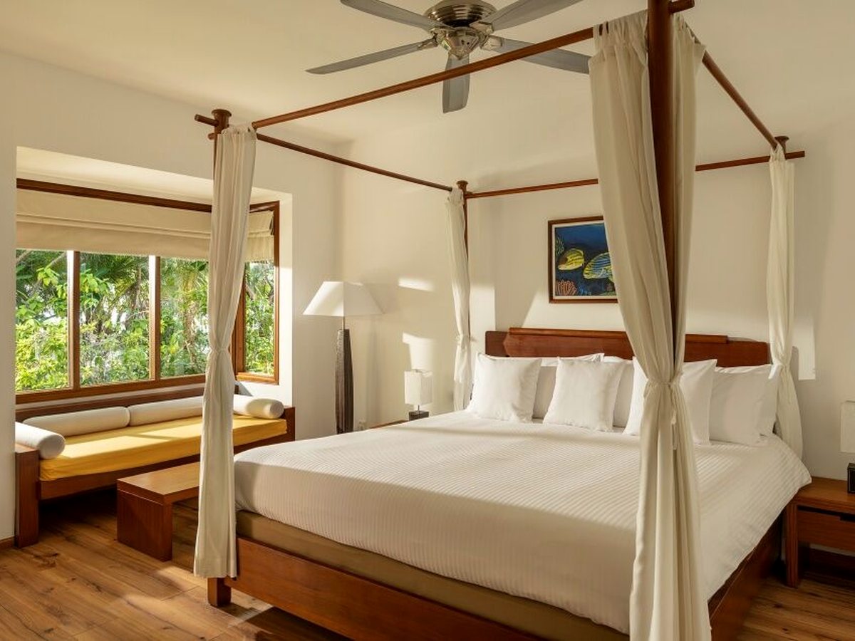 A bedroom with a wooden four-poster bed draped in white curtains, a ceiling fan above and a yellow-cushioned daybed under a window looking into the garden.