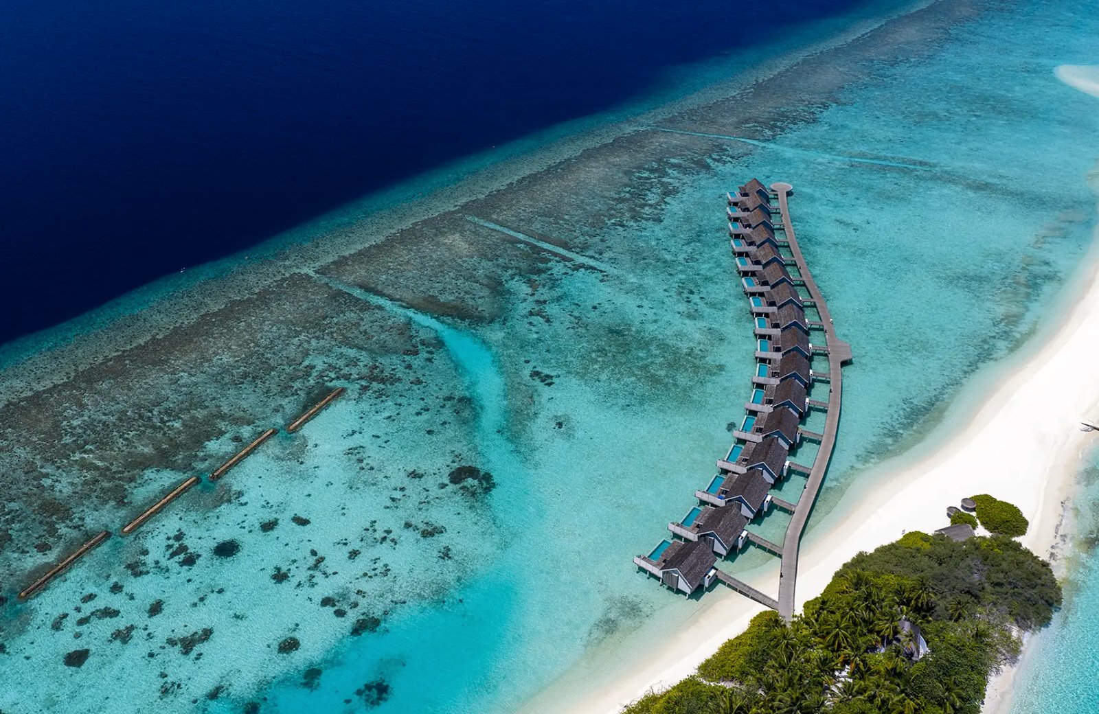 NIVA Kuramathi in the Maldives from the air: a long curving jetty of thatched overwater villas standing in a turquoise lagoon over the reef, with the island's white sand and palms at one end.