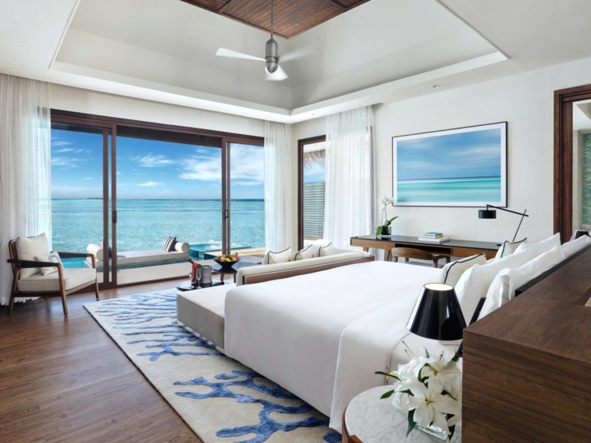 A bright villa bedroom with a king bed, blue coral-patterned rug, ceiling fan and writing desk, with sliding glass doors open to a deck, a pool and the turquoise lagoon beyond.