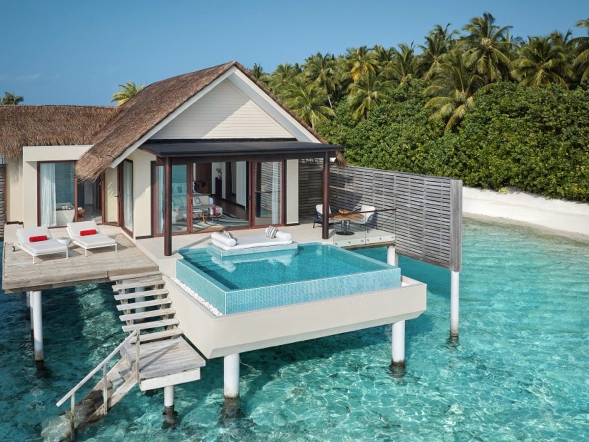 A thatched overwater villa on stilts with a private plunge pool and sun loungers on its deck, and a timber staircase leading down into clear shallow turquoise water.