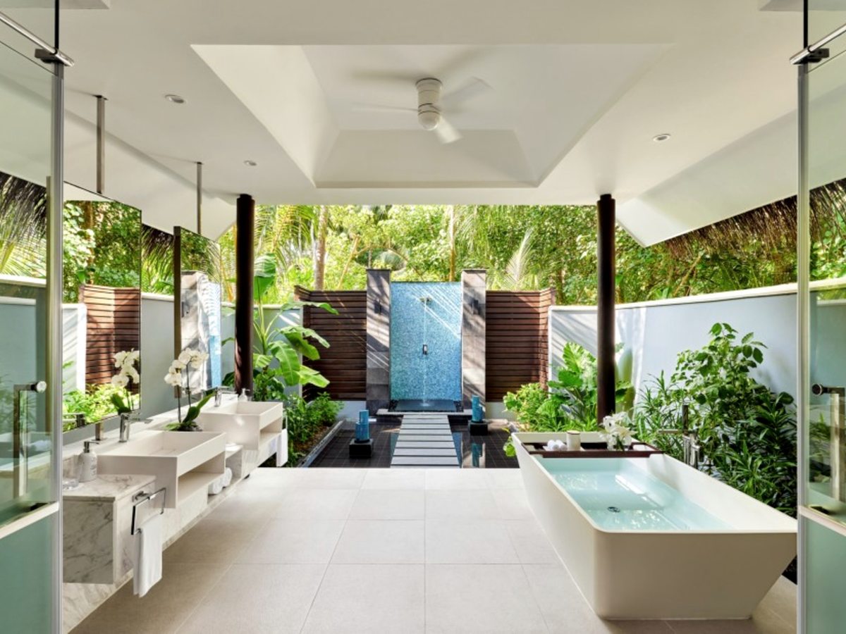 An indoor-outdoor bathroom with twin marble basins and a freestanding bath, opening onto a walled garden courtyard with a mosaic-tiled rain shower and tropical planting.