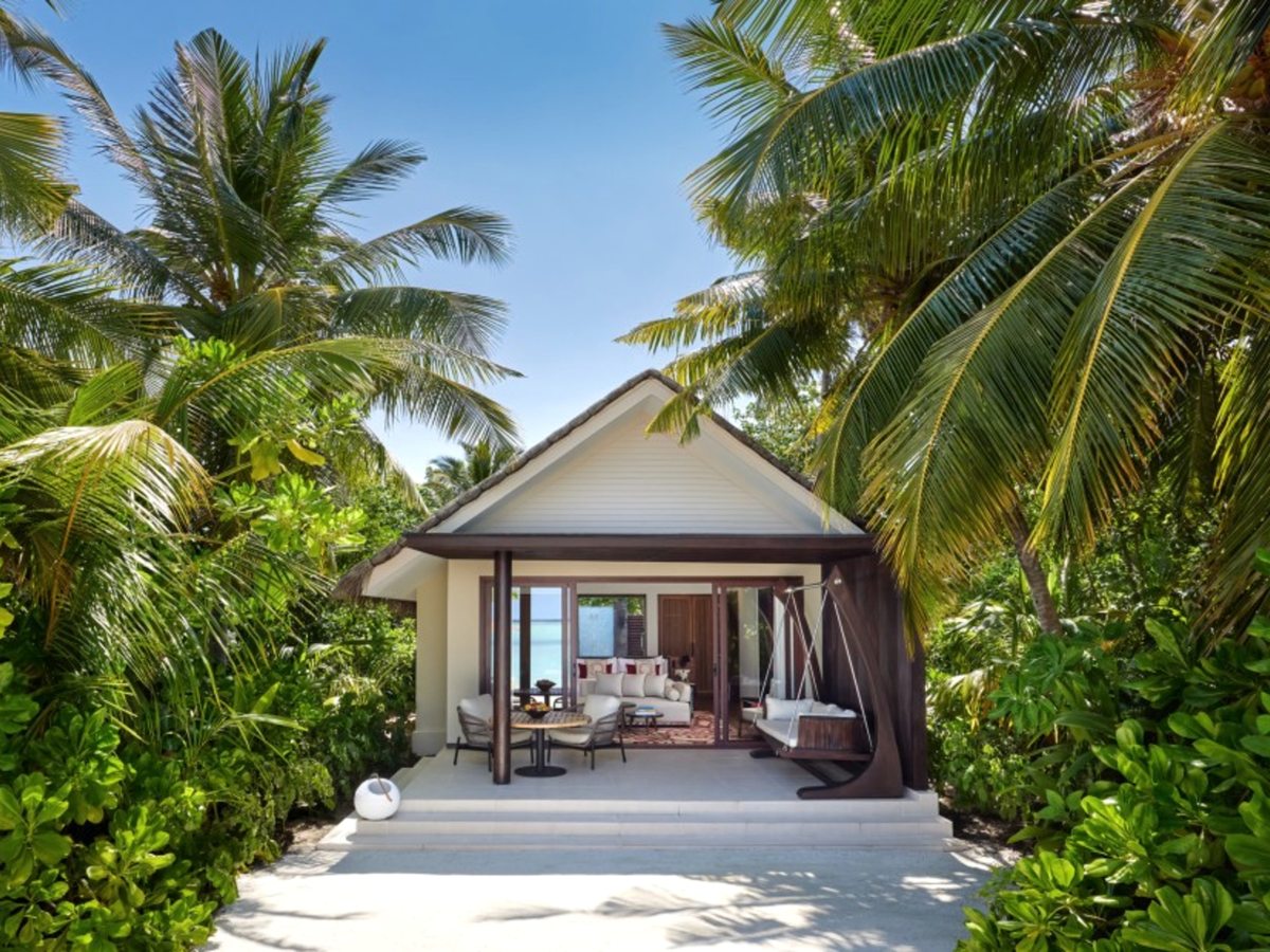 A thatched beach villa with an open-fronted living area and a hanging swing seat, framed by coconut palms and dense green foliage at the end of a white sandy path.