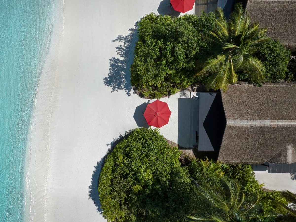 Aerial view looking straight down on a wide white-sand beach edged with turquoise shallows, with red parasols and thatched villa roofs among dense green planting along the shore.