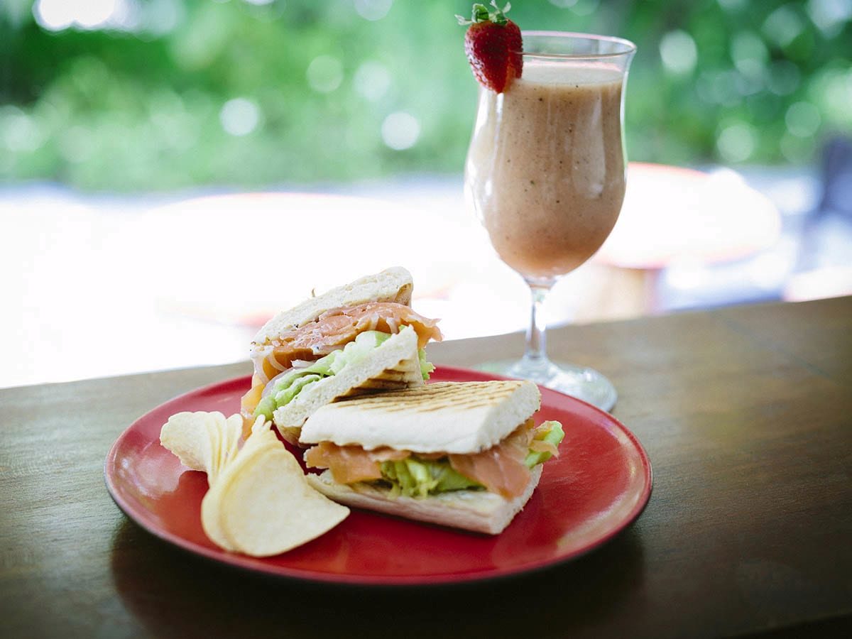 A toasted smoked salmon panini with salad and crisps on a red plate, beside a tall glass of fruit smoothie topped with a strawberry, on a dark wooden table with greenery behind.