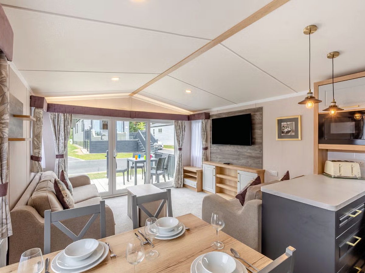 Caravan interior with a laid wooden dining table in the foreground, sofas, a wall-mounted television and patio doors opening onto decking.