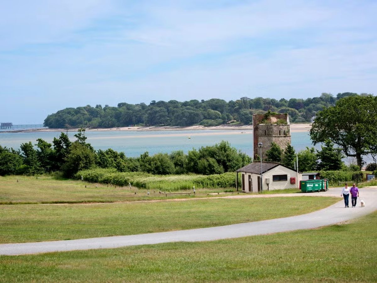 Open grass and a tarmac path running down towards the shore, with a stone tower, a small building and a sandy spit across the water.