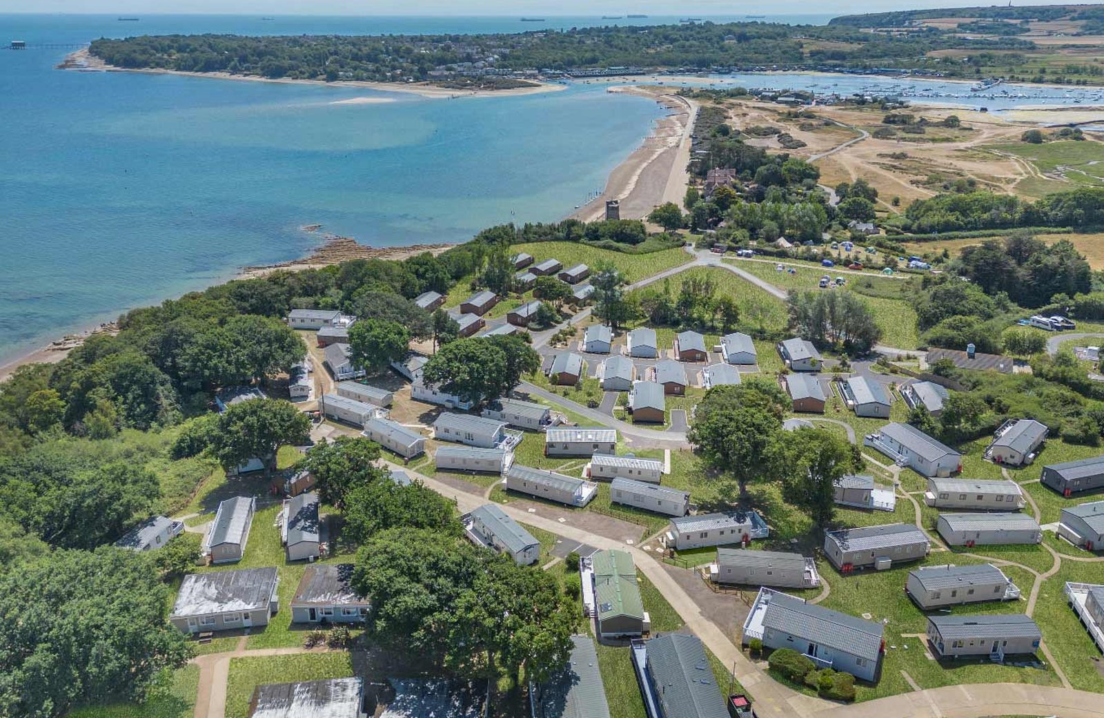 Nodes Point Holiday Park from the air: static caravans and lodges spread across a wooded slope above a long sandy beach near St Helens on the Isle of Wight, with the harbour and its moorings beyond.