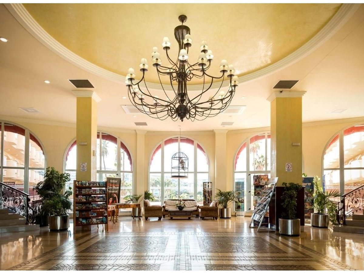 Double-height lobby with a large chandelier under a domed ceiling, tall arched windows, a polished marble floor, potted plants and seating around the edges.