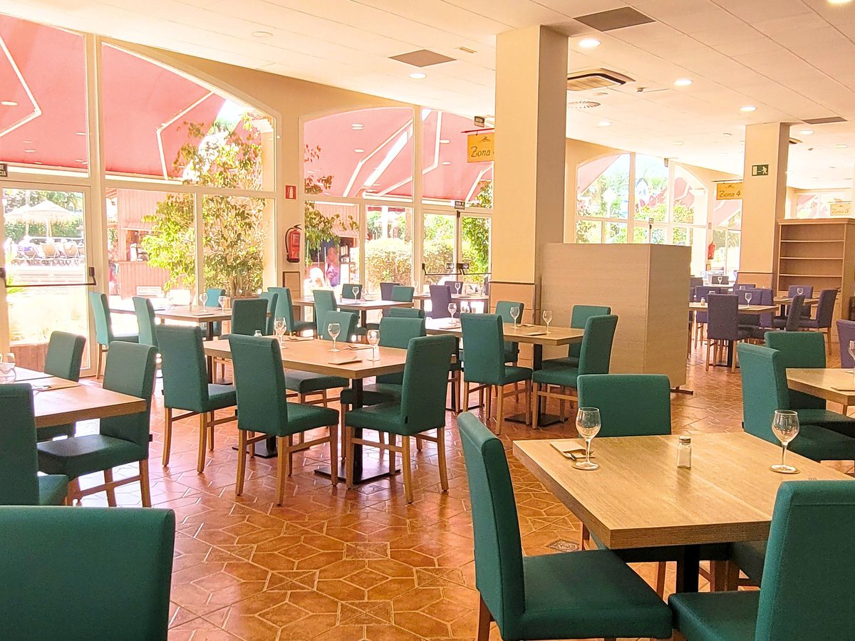 Buffet restaurant dining room with light wooden tables, teal and navy upholstered chairs, a terracotta tiled floor and tall windows onto a terrace shaded by red awnings.