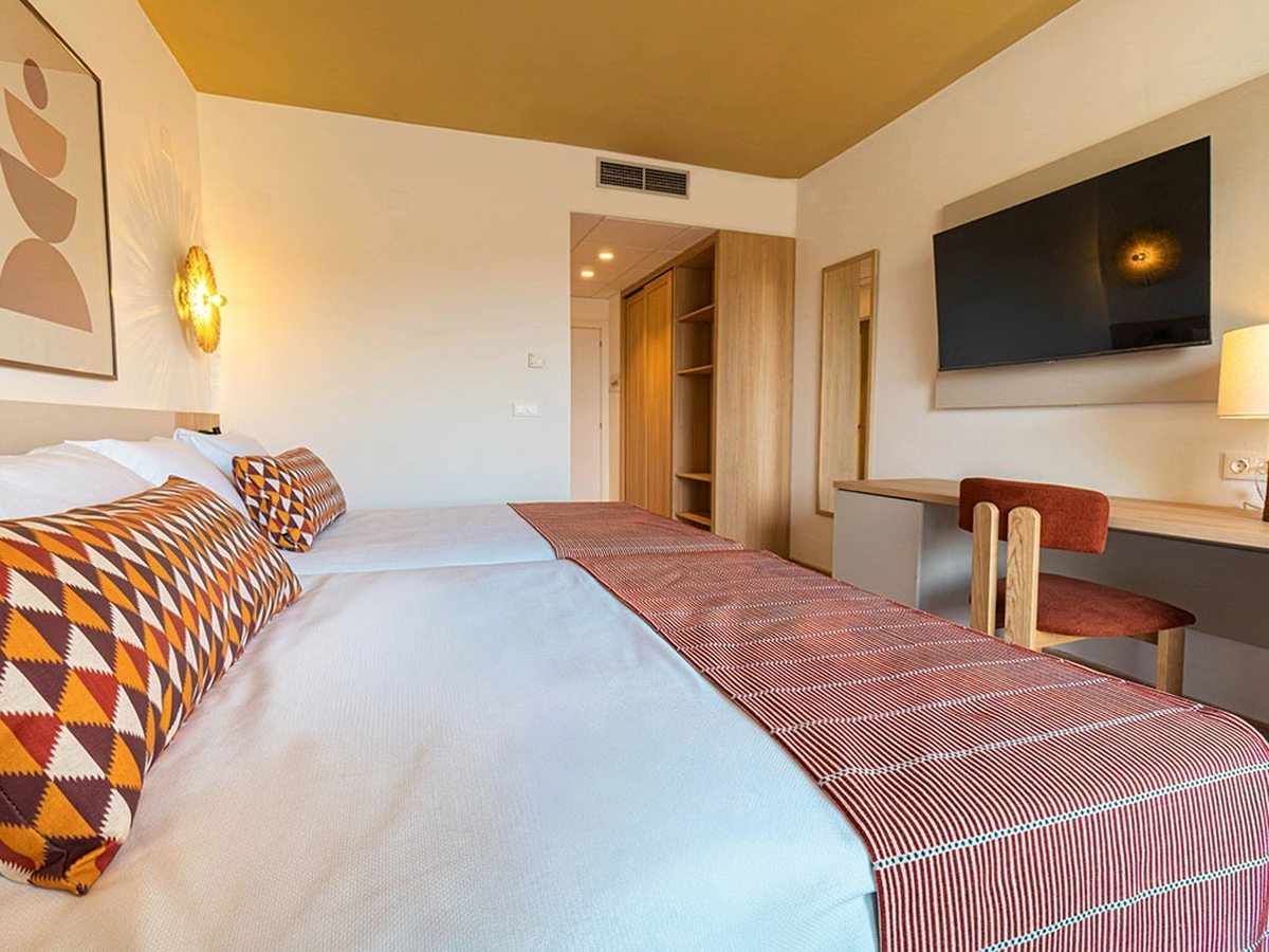 Twin room with a wall-mounted television, a pale wooden desk and rust-coloured chair, an open wardrobe with shelving, and beds dressed in white with terracotta runners.