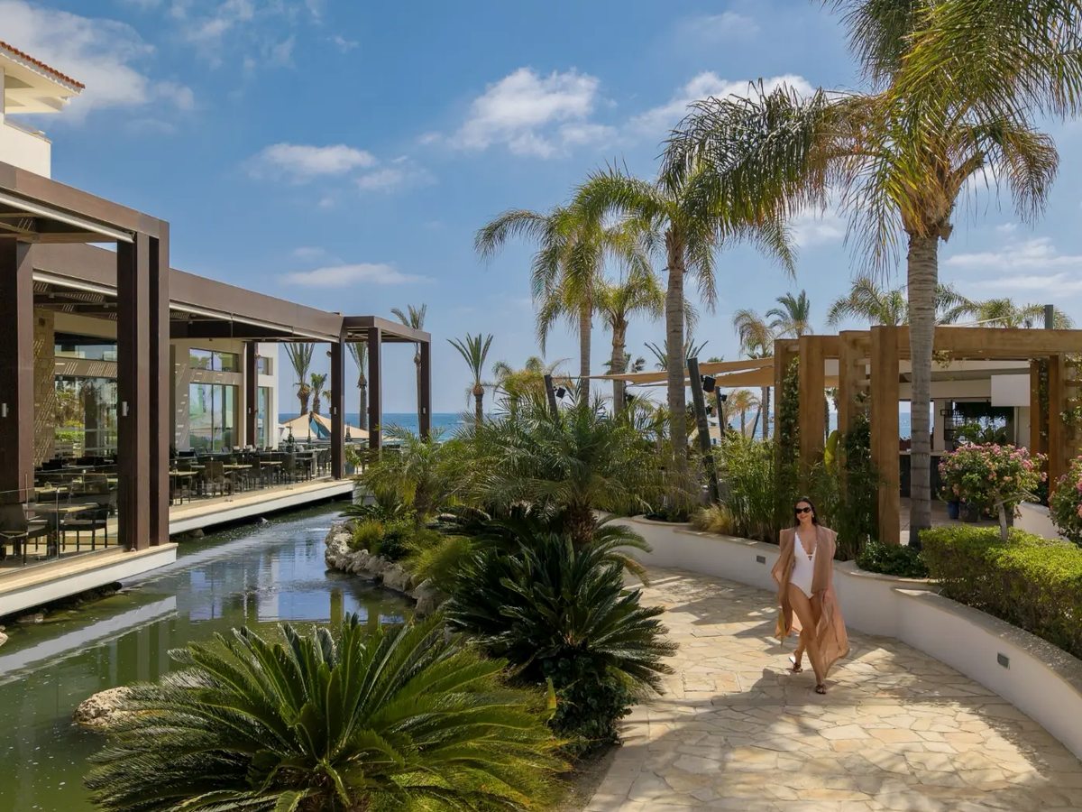 Paved walkway running alongside a long ornamental water channel, with cycads, palms and flowering shrubs, a covered dining terrace to one side and the sea in the distance.