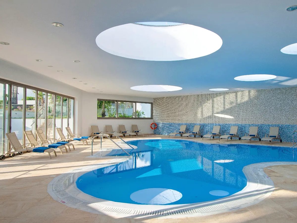 Indoor pool with a curved shallow end, round skylights in the white ceiling, mosaic tiling and rows of loungers along the glazed wall.