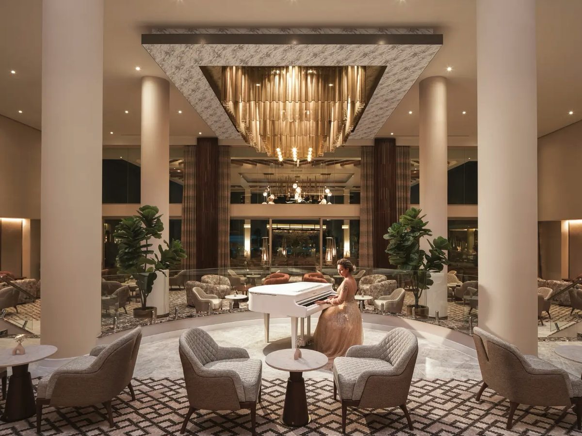 Tall lobby atrium with pale columns, a wide gold-fringed chandelier, grey armchairs on a patterned floor and a pianist at a white grand piano in the middle.