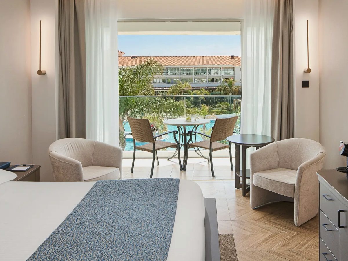 Room with a bed in the foreground, two armchairs and a balcony with a table and chairs looking over a pool and the resort buildings.