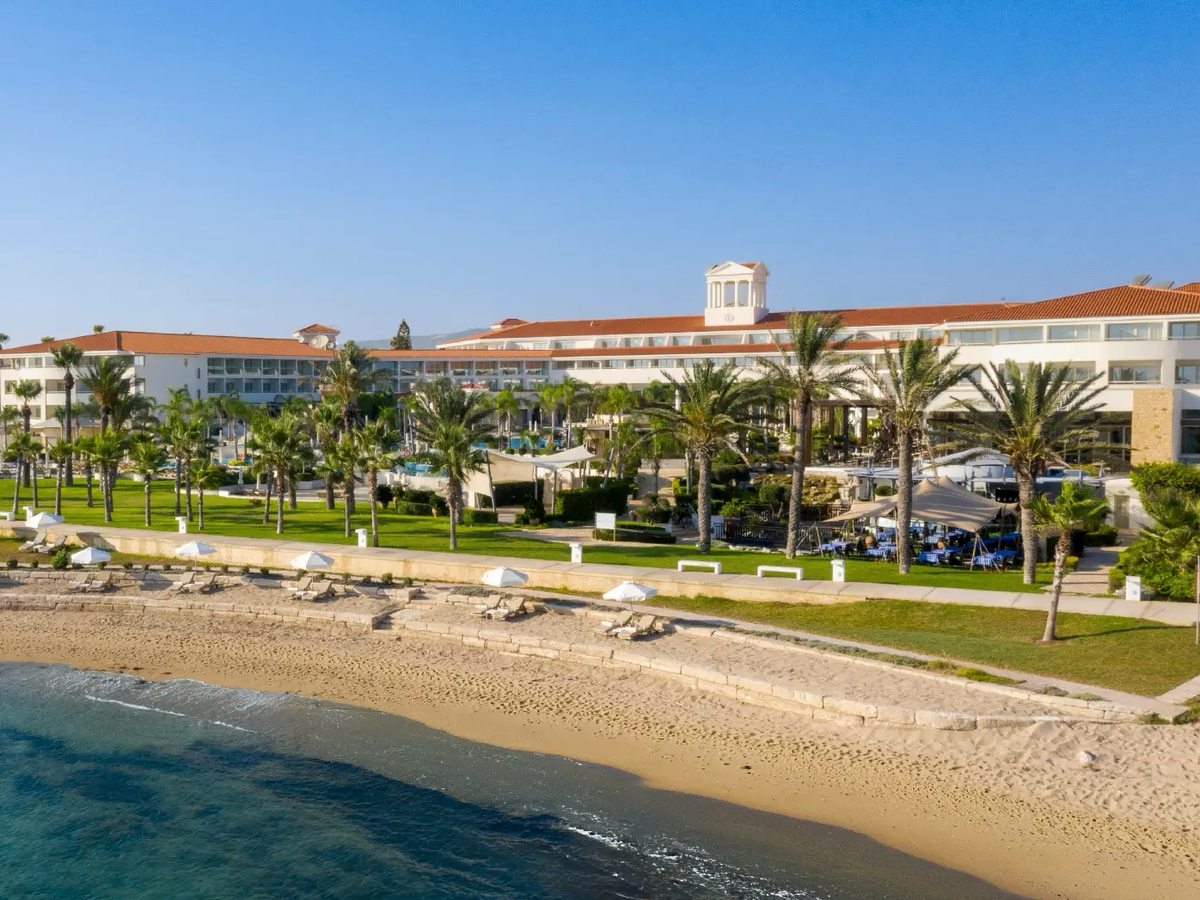 Sandy beach with rocks at the waterline, backed by the resort's lawns, palms and long white buildings with red-tiled roofs.