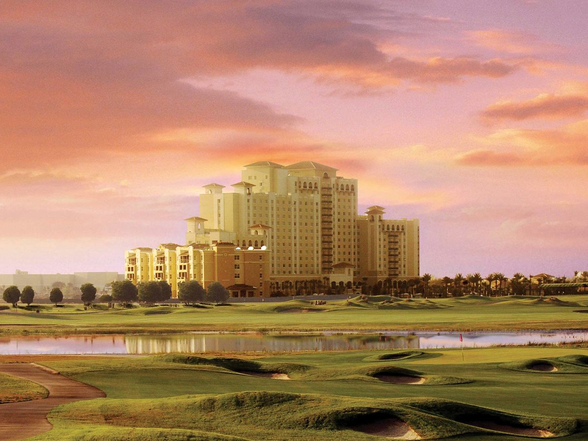 A tall resort building beyond a golf course of fairways, bunkers and a lake, under a pink and orange sunset sky.