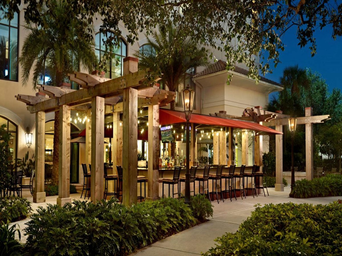 An open-air poolside bar with a timber pergola and red canopy, bar stools at the counter and tropical planting, photographed at dusk.
