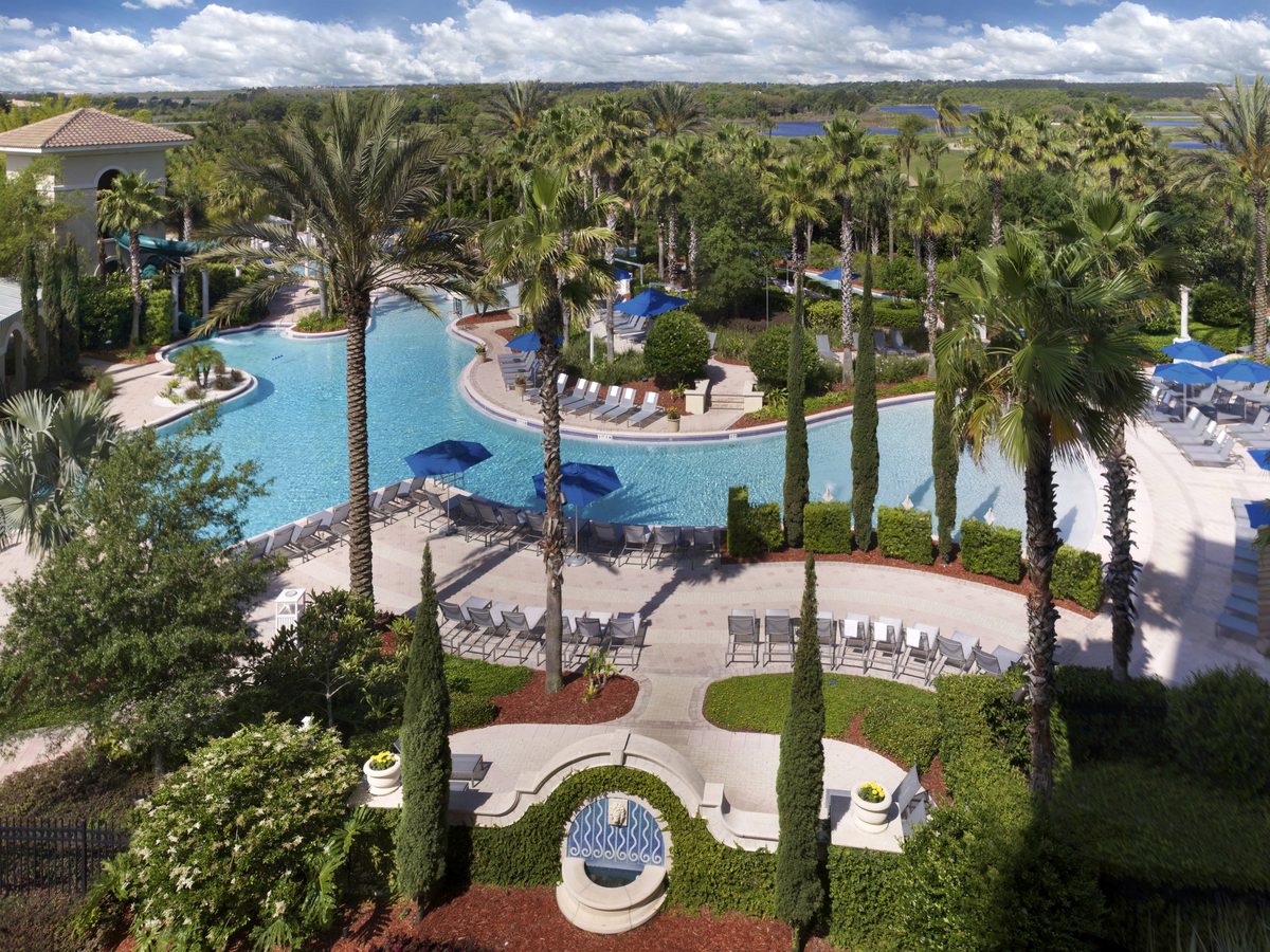 A freeform lagoon-style pool seen from a terrace, with a landscaped island, palm trees, sun loungers and blue parasols beside a hotel colonnade.