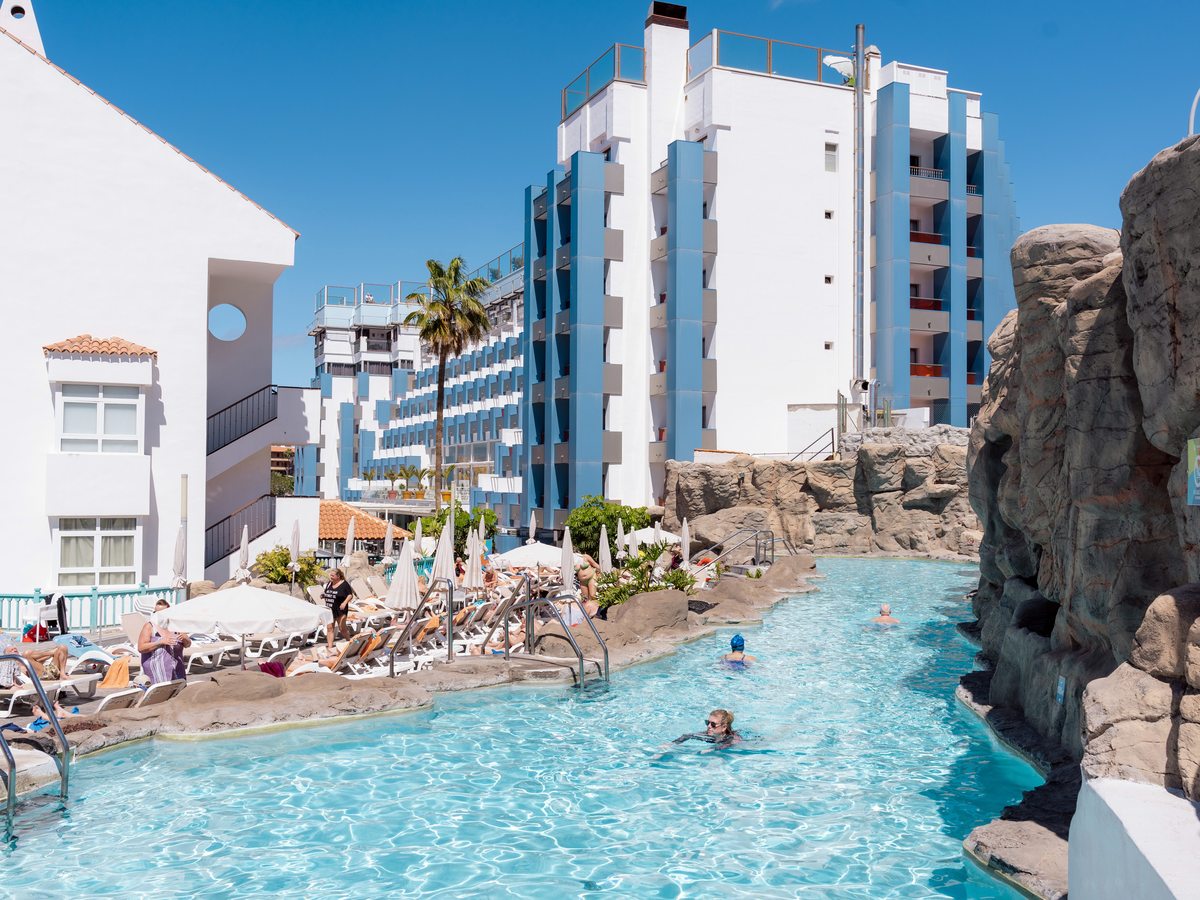 Long pool edged with sculpted grey-brown rock, swimmers in the water, sun loungers along one side and the hotel's blue and white tower rising behind.