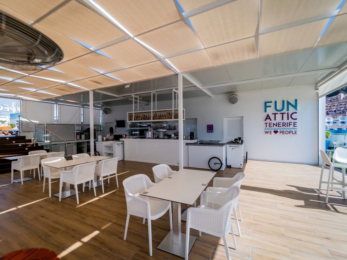 Open-sided rooftop bar with white tables and chairs on a wooden floor under a cream shade canopy, with a white service counter and a FunAttic Tenerife wall sign.