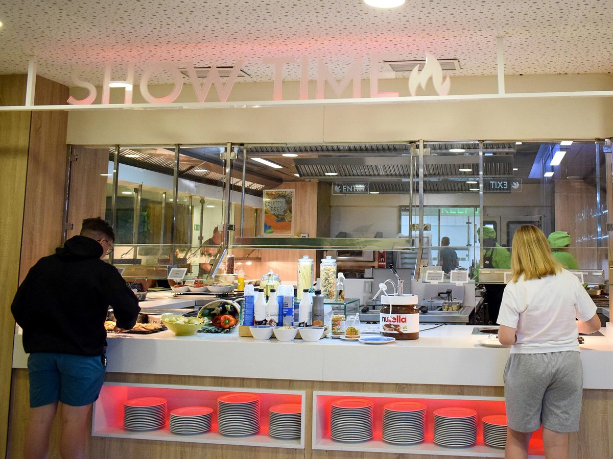 Guests standing at a long white counter under a lit SHOW TIME sign, with chefs working in the open kitchen behind a glass screen and stacks of plates in red-lit recesses below.