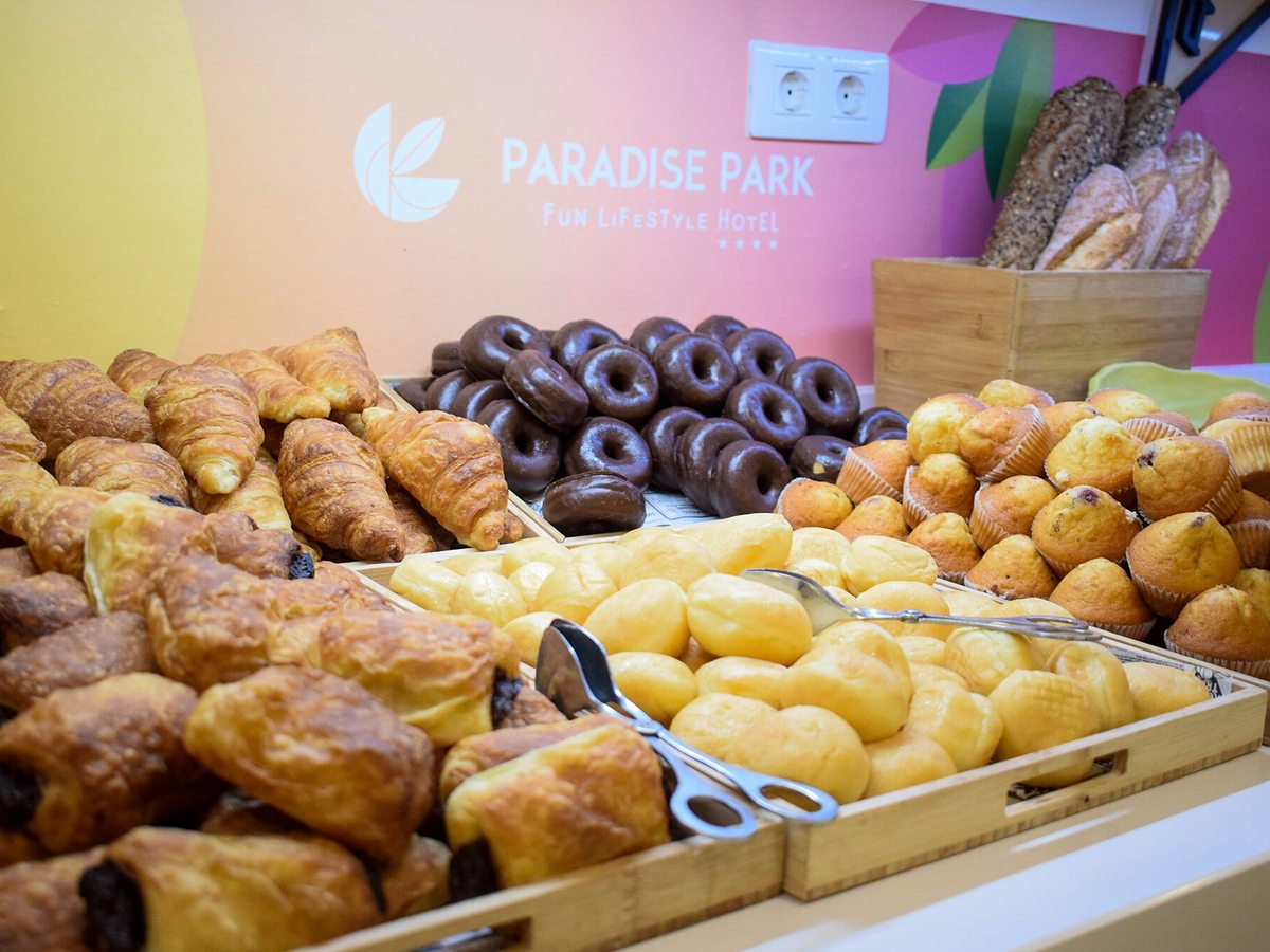Wooden trays of croissants, pains au chocolat, chocolate-topped doughnuts, muffins and soft rolls in front of a pink and orange Paradise Park wall.