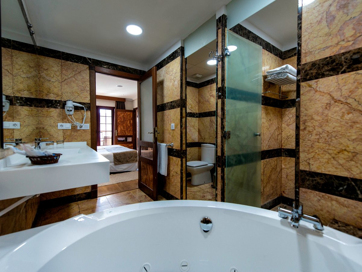 Marble-lined bathroom with a large round bath in the foreground, a glass-enclosed shower, a long white washbasin and a doorway through to the bedroom.