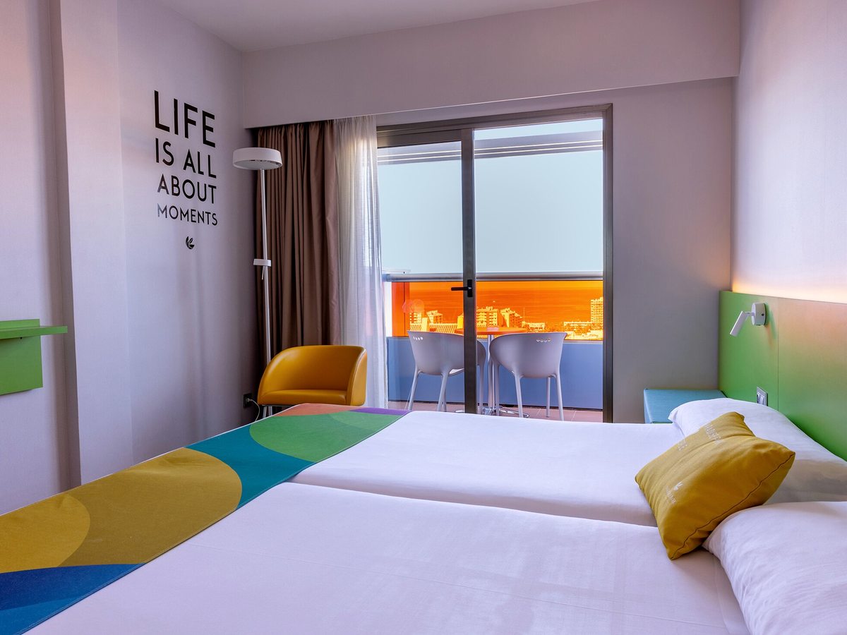 Twin beds beside sliding glass doors onto a balcony with a table and two chairs, an orange glazed screen and the town's rooftops beyond.