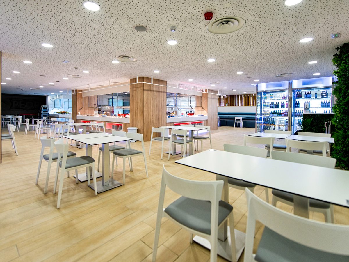 Bright buffet restaurant with a pale wood floor, white tables and chairs, wood-clad serving counters with open kitchens behind and glass-fronted drinks fridges to one side.