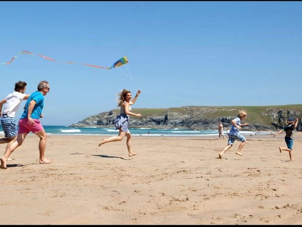 Adults and children running barefoot across firm golden sand flying a small rainbow kite, with breaking waves and a green-topped headland behind them.