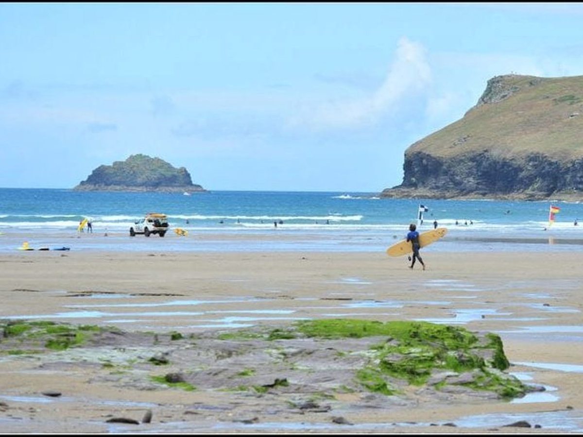 A wide beach at low tide with weed-covered rock and shallow pools in the foreground, an RNLI lifeguard vehicle and red and yellow flags at the water's edge, a surfer carrying a yellow longboard, and a green-topped headland to the right.