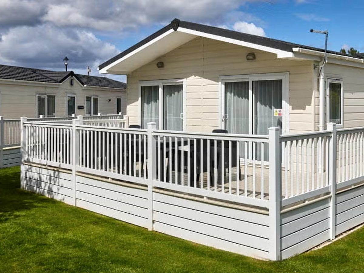 A cream-clad holiday lodge with an apex roof and patio doors, fronted by a railed veranda with a table and chairs, on a grass pitch with other units behind.