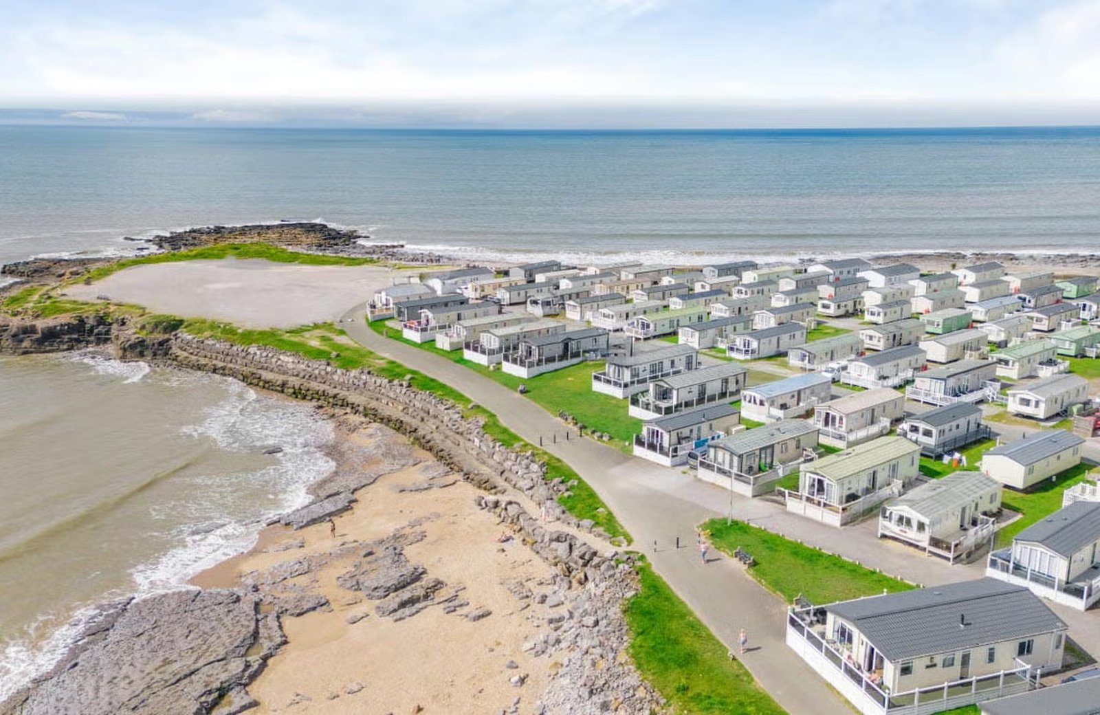 Rows of static caravans on grass pitches beside a sandy, rock-strewn shore and the sea, at a large holiday park on the south Wales coast.
