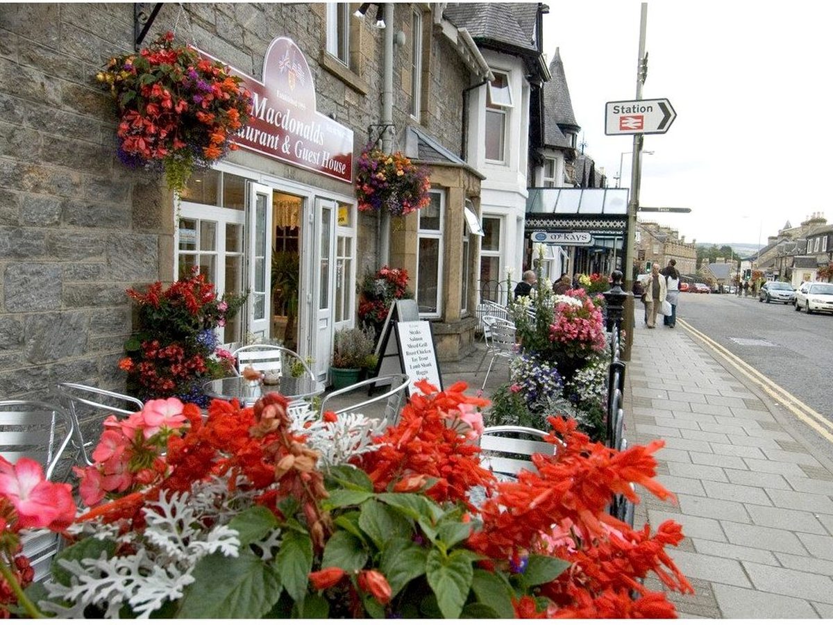 A stone-fronted hotel and restaurant on Pitlochry's high street, with pavement tables, hanging baskets and red flowers in the foreground.