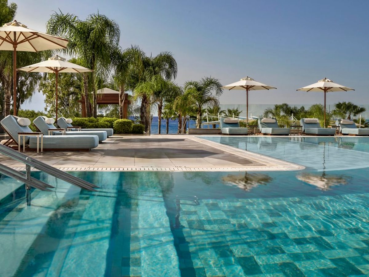 Two levels of outdoor pool separated by a stone terrace, with pale blue cushioned daybeds along the edges, palms and the Mediterranean beyond.