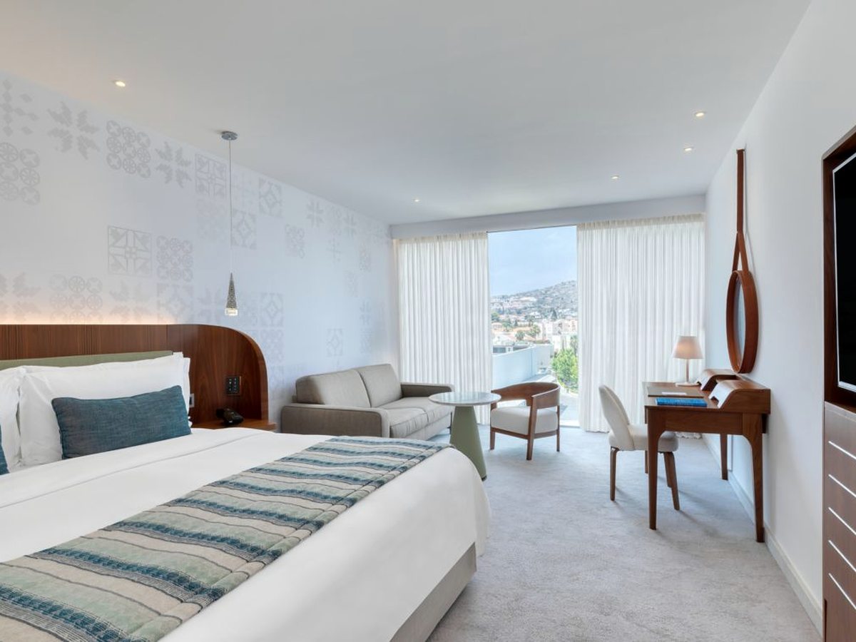 A hotel room with a king bed, a pale sofa, a writing desk and floor-to-ceiling balcony doors looking onto a green hillside and houses.