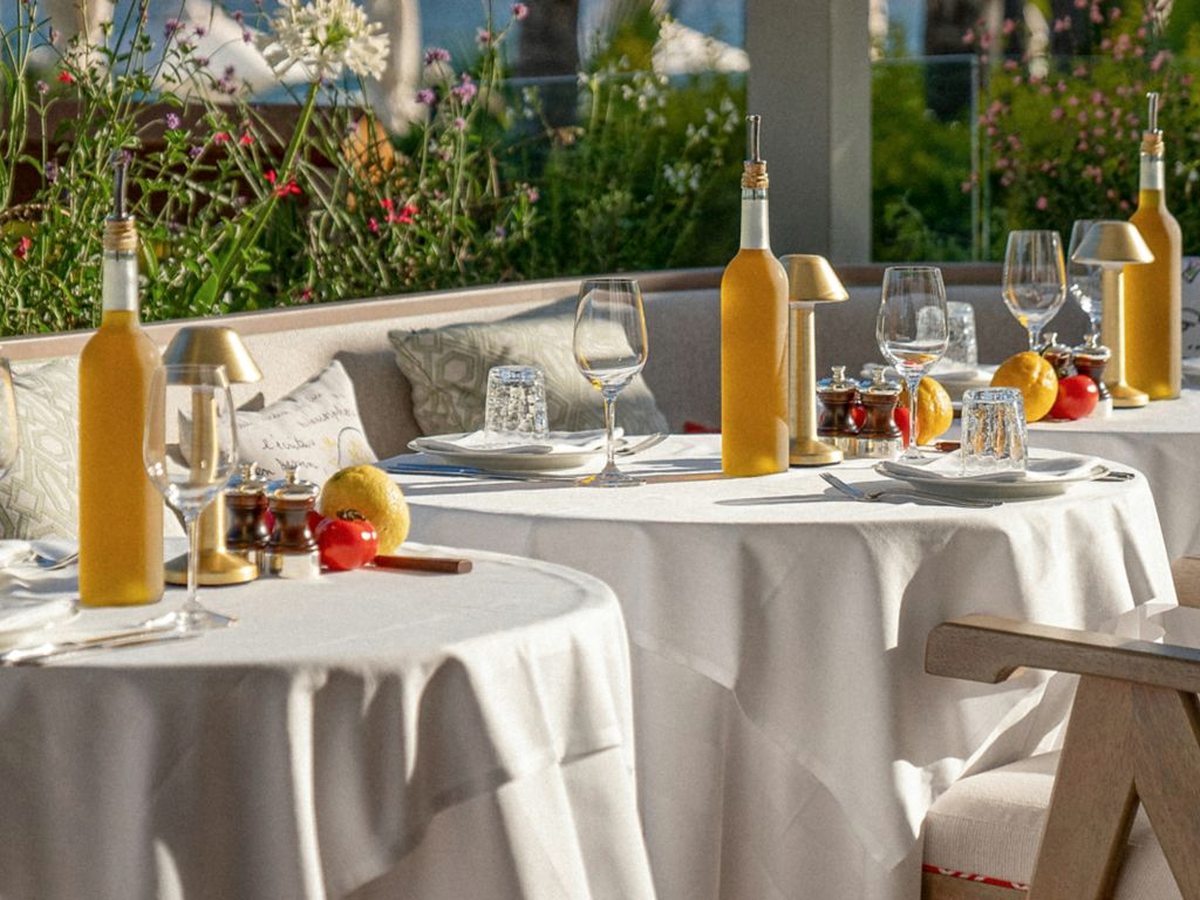 Round tables under white linen on a terrace, laid with wine glasses, olive-oil bottles and small brass lamps, backed by flowering shrubs.