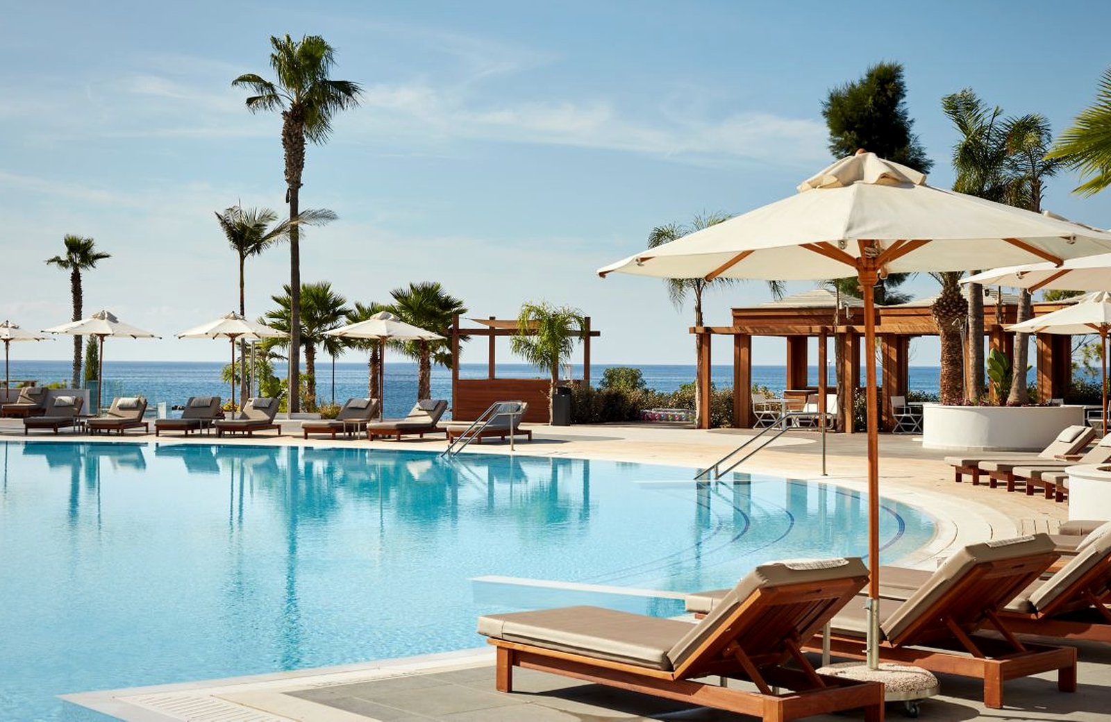 A large curved outdoor pool at Parklane, edged with wooden sun loungers and cream parasols, with palms, timber cabanas and the Mediterranean behind.