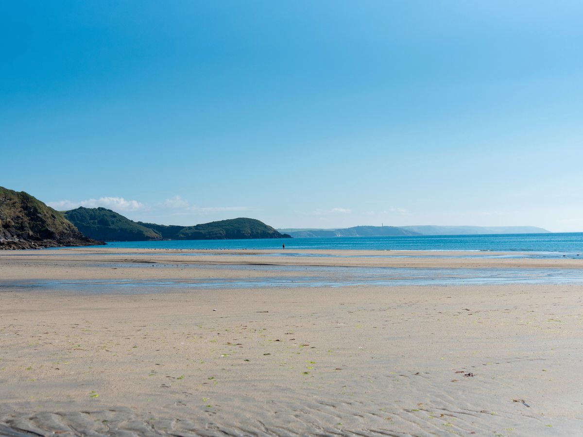 A wide flat sandy beach at low tide with shallow pools of water, calm blue sea and a wooded headland closing the bay.