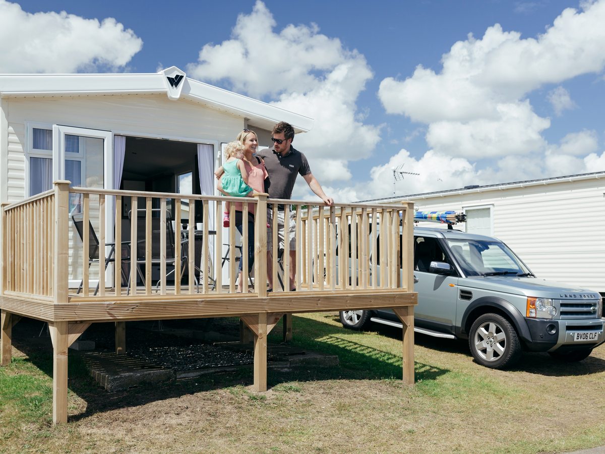 A cream static holiday home with patio doors opening onto a raised timber sundeck, a family standing at the rail and a car parked on the grass beside it.